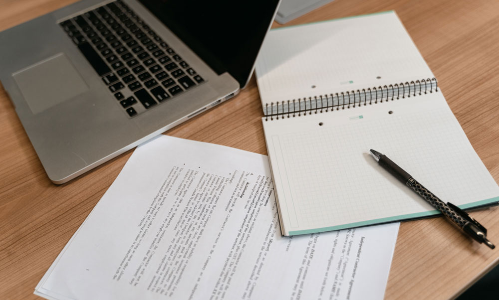 Laptop, spiral notebook, pen, and printed documents on a wooden desk, ready for study or work import.